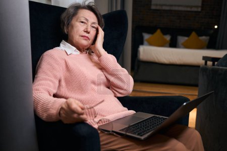 Foto de Agradable señora de edad avanzada se encuentra con un ordenador portátil en una silla acogedora, ella está en ropa casual cómoda - Imagen libre de derechos