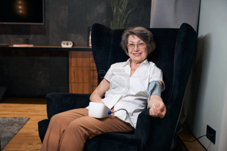 Foto de Señora anciana optimista se sienta en una silla y mide su presión arterial con un esfigmomanómetro automático - Imagen libre de derechos