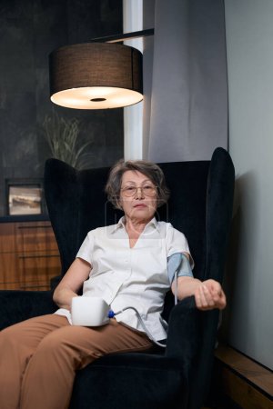 Téléchargez les photos : Calme vieille femme assise dans un fauteuil et mesure sa pression artérielle avec un sphygmomanomètre automatique - en image libre de droit