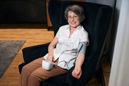 Téléchargez les photos : Agréable dame âgée s'assoit dans un fauteuil facile et mesure sa pression avec un tonomètre automatique - en image libre de droit