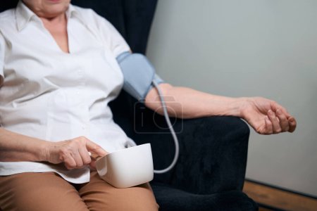 Foto de Una anciana solitaria se sienta en una silla y mide su presión arterial con un esfigmomanómetro automático - Imagen libre de derechos