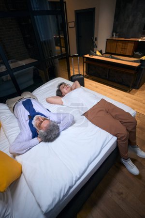 Téléchargez les photos : Fatigué de la route, un homme âgé et son compagnon se reposent sur le lit de l'hôtel, valise de voyage est debout à proximité - en image libre de droit