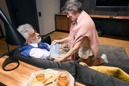 Foto de Esposa de pelo gris mide la presión de su marido con un tonómetro, la pareja se encuentra en un cómodo sofá - Imagen libre de derechos