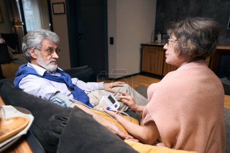 Foto de La esposa preocupada mide la presión arterial al marido viejo, pareja se sienta en el sofá cómodo - Imagen libre de derechos