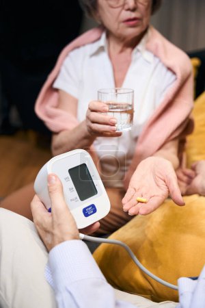 Foto de La anciana esposa le da a su marido pastillas y un vaso de agua, un hombre mide la presión arterial con un tonómetro - Imagen libre de derechos