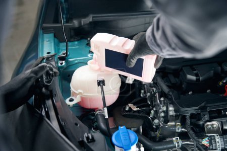 Téléchargez les photos : Mécanicien en vêtements de protection debout près de la voiture avec capot ouvert et changeant antigel - en image libre de droit
