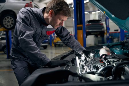 Téléchargez les photos : Mécanicien en vêtements de protection debout dans le montage des pneus et faire l'inspection du moteur de voiture - en image libre de droit