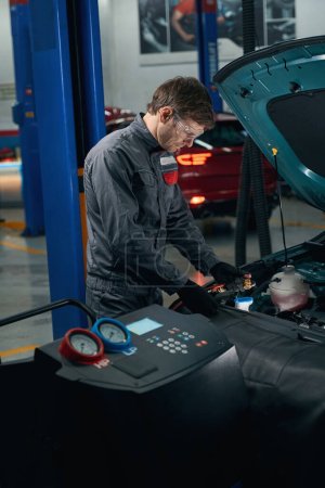 Foto de Mecánico en ropa protectora de pie en el montaje de neumáticos cerca de coche y repostaje de aire acondicionado - Imagen libre de derechos