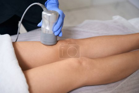 Photo for Lady lying on couch in salon, medic in protective gloves doing RF lifting for leg - Royalty Free Image
