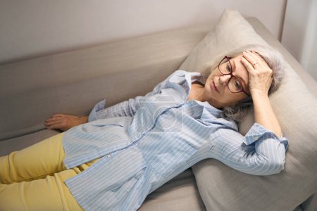 Foto de La anciana cansada yace en el sofá y sostiene la cabeza, no está bien - Imagen libre de derechos