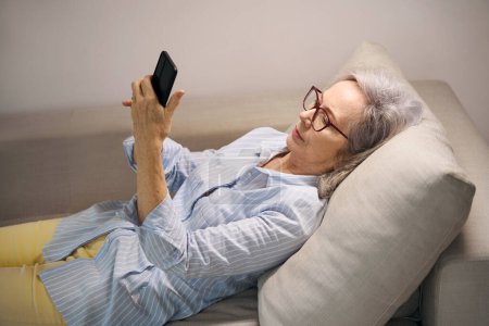 Téléchargez les photos : Femme âgée reposant à la maison regarde quelque chose sur son téléphone, elle est située sur un canapé doux - en image libre de droit