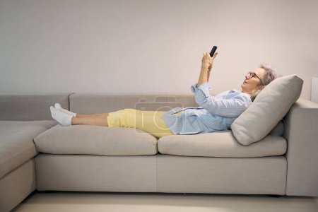 Photo for Gray-haired lady in a cozy home environment lies on the sofa, she uses the phone - Royalty Free Image