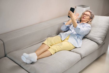 Foto de Anciana de buen humor se encuentra en el sofá en casa, ella utiliza el teléfono - Imagen libre de derechos