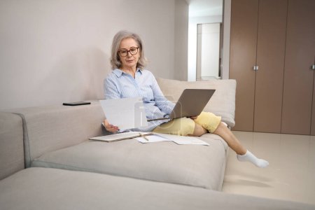 Photo for Elderly lady is sitting on a sofa with laptop on her knees, she has piece of paper in her hands - Royalty Free Image