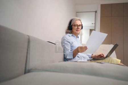 Photo for Cute grandmother is studying distance learning at home on the couch, she has a computer headset - Royalty Free Image