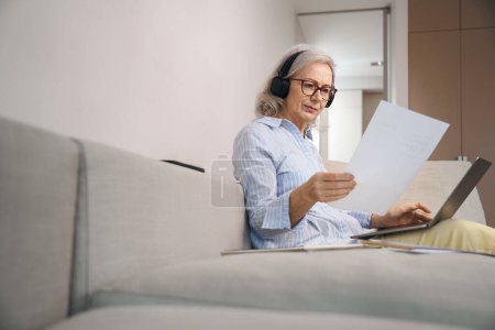 Téléchargez les photos : Femme aux cheveux gris concentré lit un résumé à la maison sur le canapé, elle a un ordinateur portable sur les genoux - en image libre de droit