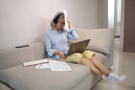 Photo for Busy elderly lady is working on a laptop at home on the couch, she is using a computer headset - Royalty Free Image