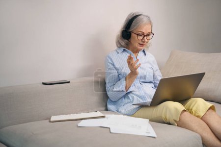 Photo for Active grandmother studying online courses at home on the couch, she has a computer headset - Royalty Free Image