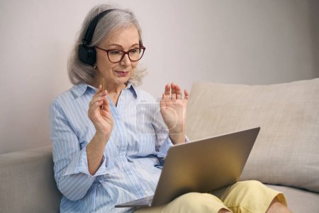 Foto de Moderna abuela en gafas y auriculares de computadora se sienta en el sofá con un ordenador portátil, ella tiene un lápiz en sus manos - Imagen libre de derechos