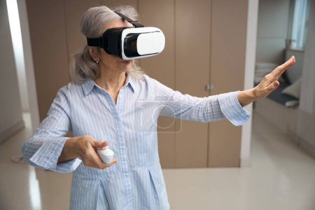 Téléchargez les photos : Femme aux cheveux gris se tient dans le salon en lunettes de réalité virtuelle, elle a un panneau de contrôle dans ses mains - en image libre de droit