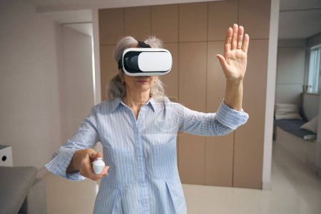 Foto de Pensionista moderna se encuentra en la sala de estar en gafas de realidad virtual, ella tiene un panel de control en sus manos - Imagen libre de derechos