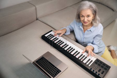 Photo for Pleasant elderly woman playing digital piano in living room, laptop standing in front of her - Royalty Free Image