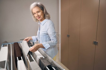 Foto de Esbelta abuela cuelga cuidadosamente la ropa después de lavarse en una secadora de habitaciones, una mujer con ropa casual - Imagen libre de derechos