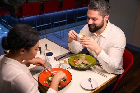 Foto de Sonriente chico bebiendo café mientras su novia comiendo ensalada en la mesa del restaurante - Imagen libre de derechos
