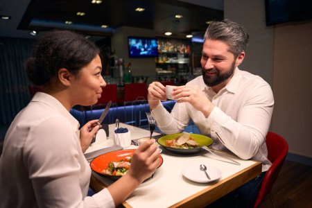 Foto de Hombre con taza de café en las manos sonriendo a su novia comiendo ensalada en la mesa del restaurante - Imagen libre de derechos