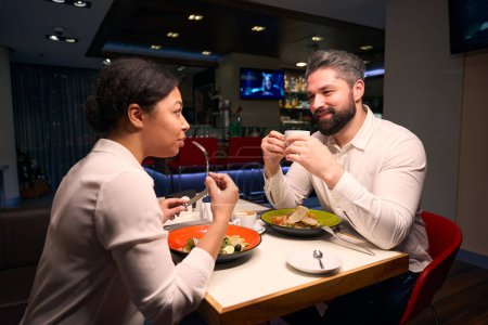 Téléchargez les photos : Guy avec une tasse de café dans les mains admirant sa compagne mangeant de la salade à la table du restaurant - en image libre de droit
