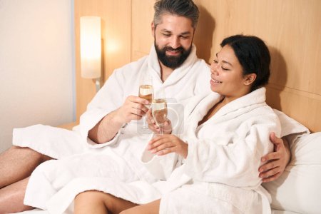 Foto de Agradable hombre y su feliz acompañante hembra tintineo copas de champán en la cama - Imagen libre de derechos
