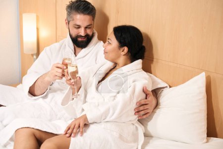 Photo for Happy young male and his female companion clinking glasses of champagne in bed - Royalty Free Image