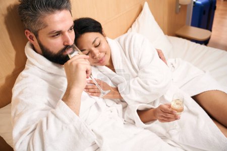 Téléchargez les photos : Pensif gars boire un verre de champagne tout en embrassant son compagnon féminin souriant au lit - en image libre de droit