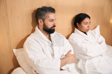Téléchargez les photos : Homme sérieux et sa compagne triste vêtue de peignoirs assis au lit - en image libre de droit