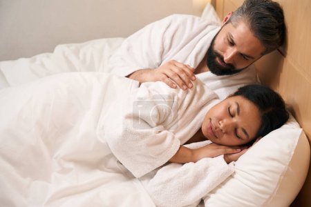 Téléchargez les photos : Soins jeune caucasien mâle étreignant son sommeil afro-américain femelle compagnon dans lit - en image libre de droit