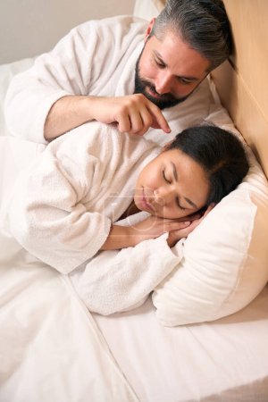 Photo for Playful man lying in bed while touching his female companion ear with index finger - Royalty Free Image