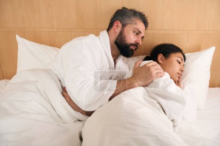 Foto de Cuidando joven macho acostado en la cama mientras tocaba su deprimido hombro de novia - Imagen libre de derechos