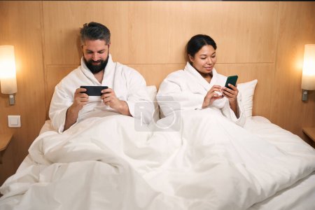 Foto de Hombre y su complacida compañera acostada en la cama con teléfonos móviles en las manos - Imagen libre de derechos