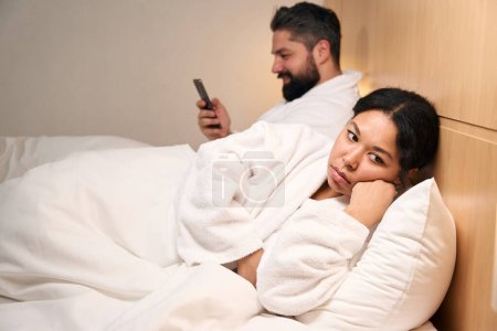 Téléchargez les photos : Triste jeune femme couchée au lit à côté de son compagnon souriant avec téléphone portable - en image libre de droit