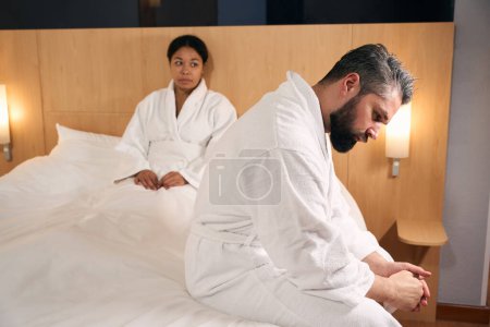 Téléchargez les photos : Triste femme regardant son petit ami bouleversé assis sur le bord du lit - en image libre de droit