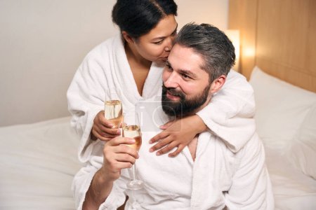 Foto de Mujer alegre con copa de champán abrazando y besando novio feliz en la cama en habitación de hotel - Imagen libre de derechos