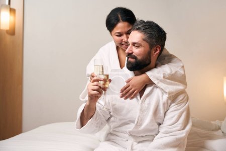 Foto de Mujer sonriente abrazo complacido hombre en la cama mientras tintineo copas de champán - Imagen libre de derechos