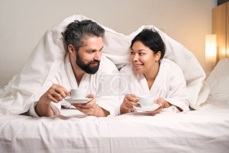 Foto de Romántica pareja joven con copas en las manos tendidas debajo de la manta en la cama - Imagen libre de derechos