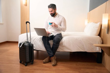 Joyous man with credit card in hand seated on bed typing on portable computer