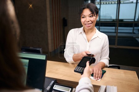 Téléchargez les photos : Carte de crédit touchante touristique féminine souriante contre terminal POS dans la main de la réceptionniste - en image libre de droit