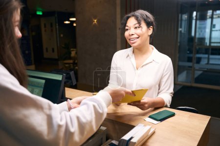 Téléchargez les photos : Jeune femme souriante recevant un formulaire d'inscription de la réceptionniste féminine à la réception - en image libre de droit