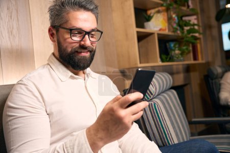 Téléchargez les photos : Portrait de jeune homme souriant assis sur un canapé regardant l'écran du smartphone - en image libre de droit