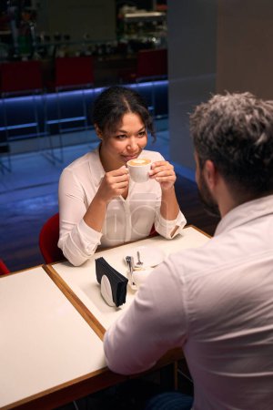 Téléchargez les photos : Jeune femme avec tasse en céramique dans les mains regardant son compagnon assis à la table de café - en image libre de droit