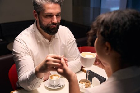 Téléchargez les photos : Romantique gars et sa compagne avec une tasse de cappuccino tenant la main à la table du café - en image libre de droit
