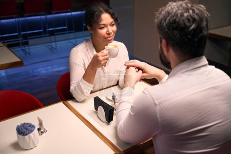 Téléchargez les photos : Souriant jeune femme heureuse avec tasse de cappuccino et homme tenant la main à la table du café - en image libre de droit